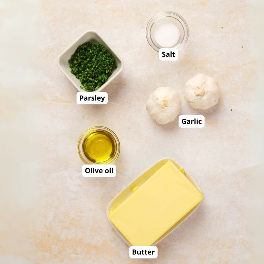 Ingredients on a surface: a bowl of chopped parsley, a pinch of salt, two garlic bulbs, a small dish of olive oil, and a block of butter.