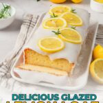 A glazed lemon loaf cake topped with lemon slices is displayed on a white platter, with fresh lemons and rosemary nearby.