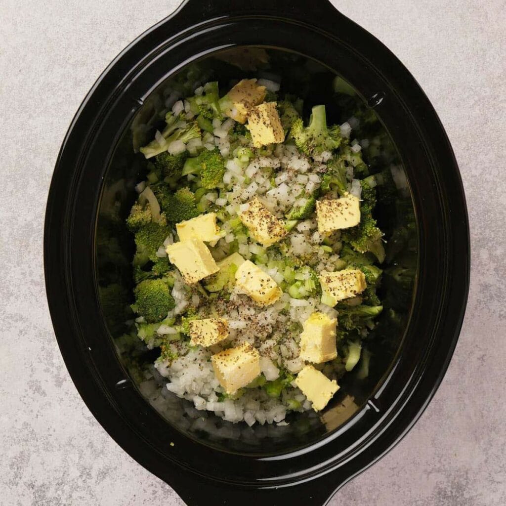 A slow cooker filled with chopped broccoli, diced onions, cubes of butter, and sprinkled with black pepper on a gray surface.
