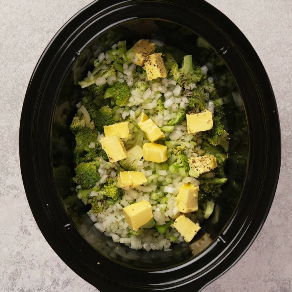 Slow cooker filled with chopped broccoli, onions, and butter cubes.