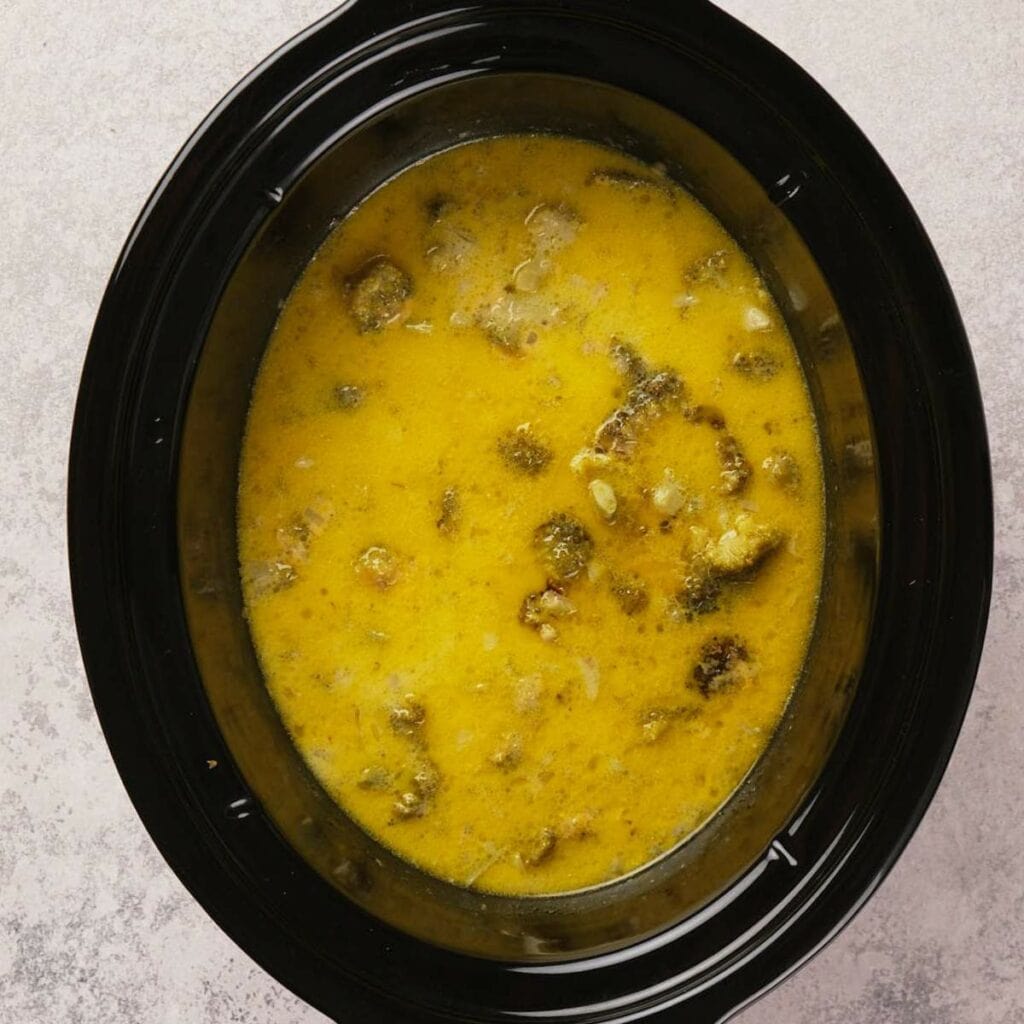 Yellow curry with visible chunks of meat and vegetables in a black slow cooker on a light gray background.