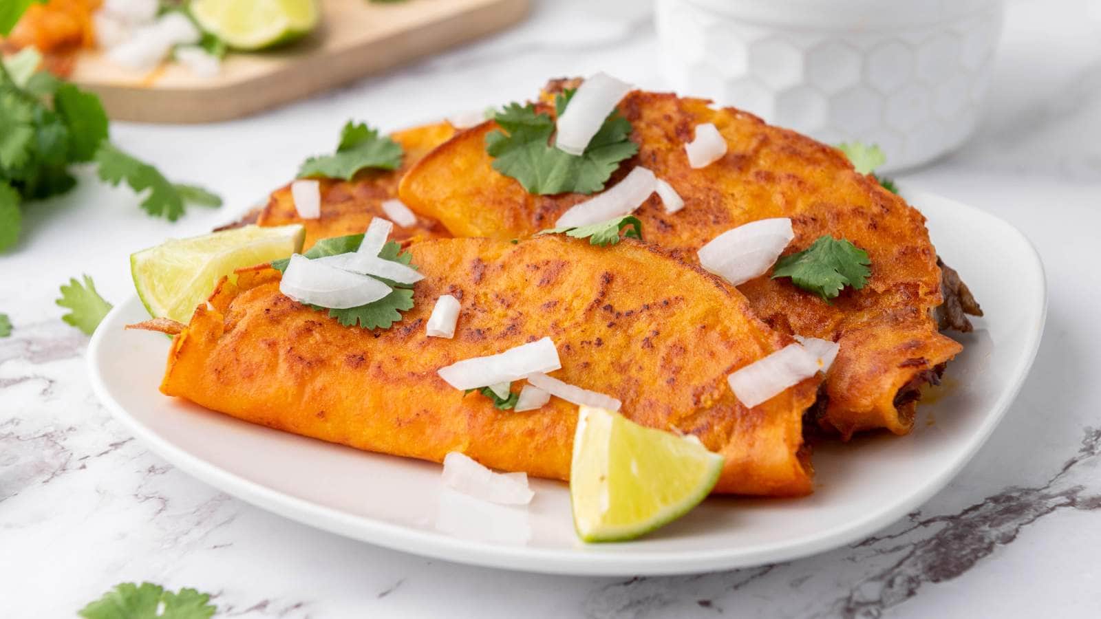 Quesabirria tacos garnished with chopped onions, cilantro, and lime wedges are served on a white plate.