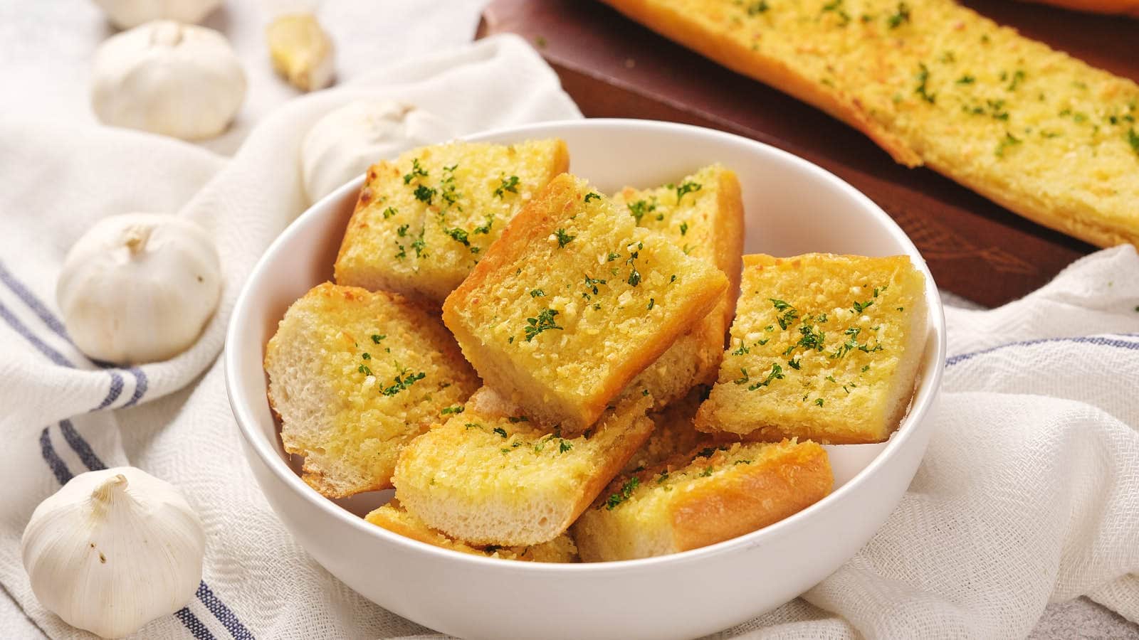 A bowl of sliced roasted garlic bread topped with herbs, surrounded by whole garlic bulbs and a striped cloth.