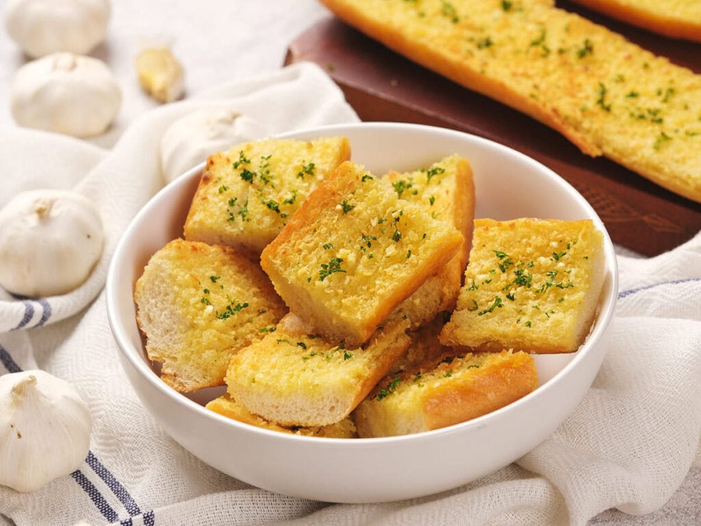 A bowl of sliced roasted garlic bread garnished with herbs, surrounded by garlic cloves.