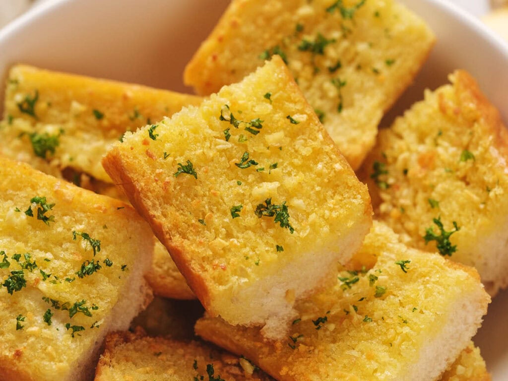 Close-up of a bowl filled with slices of roasted garlic bread, topped with chopped parsley.