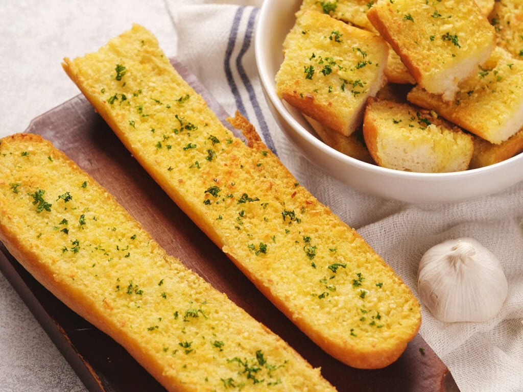 Two slices of roasted garlic bread on a wooden board and a bowl of garlic bread cubes.