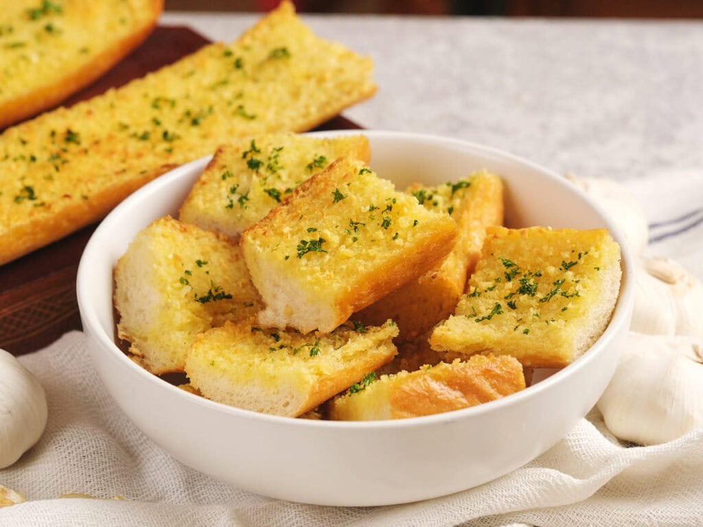 A white bowl filled with square pieces of roasted garlic bread garnished with herbs on a table.