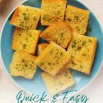 A blue plate with several pieces of roasted garlic bread garnished with herbs, placed on a light surface.