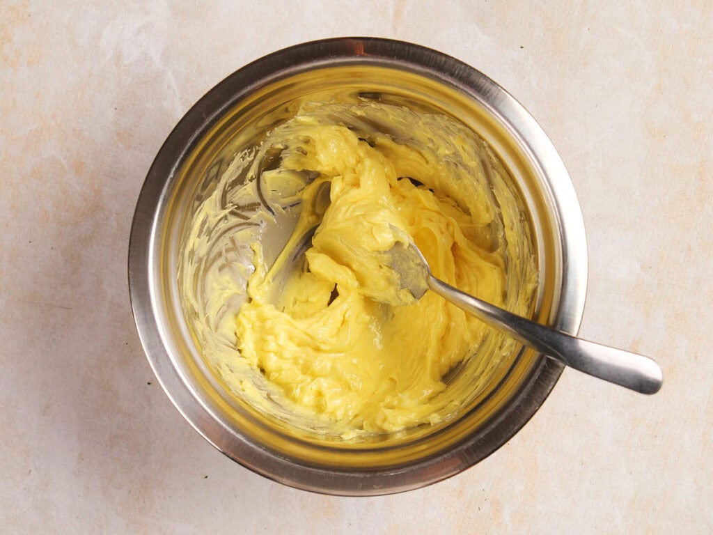A mixing bowl containing creamy yellow butter with a metal spoon inside, placed on a light-colored surface.