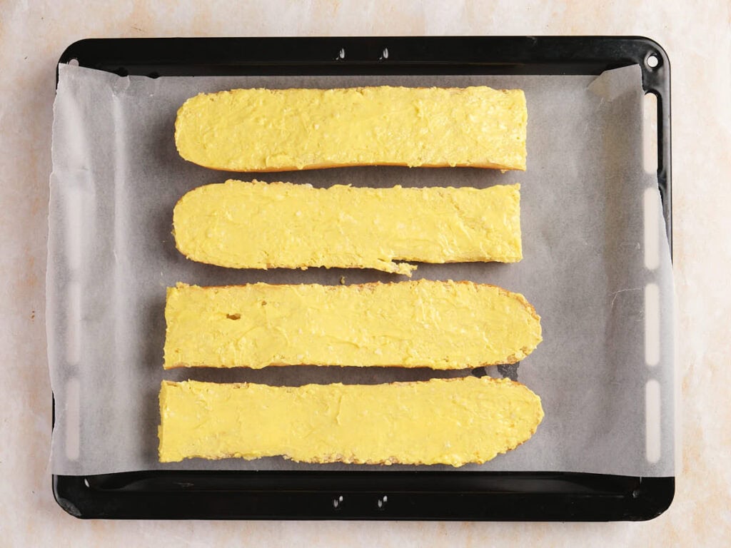 Four slices of bread spread with a yellow butter mixture are placed on a baking tray lined with parchment paper.