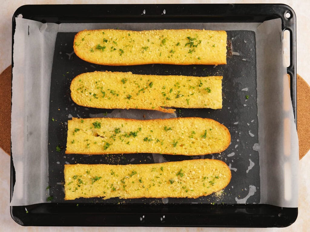 Four slices of garlic bread with parsley and seasoning on a parchment-lined baking tray.
