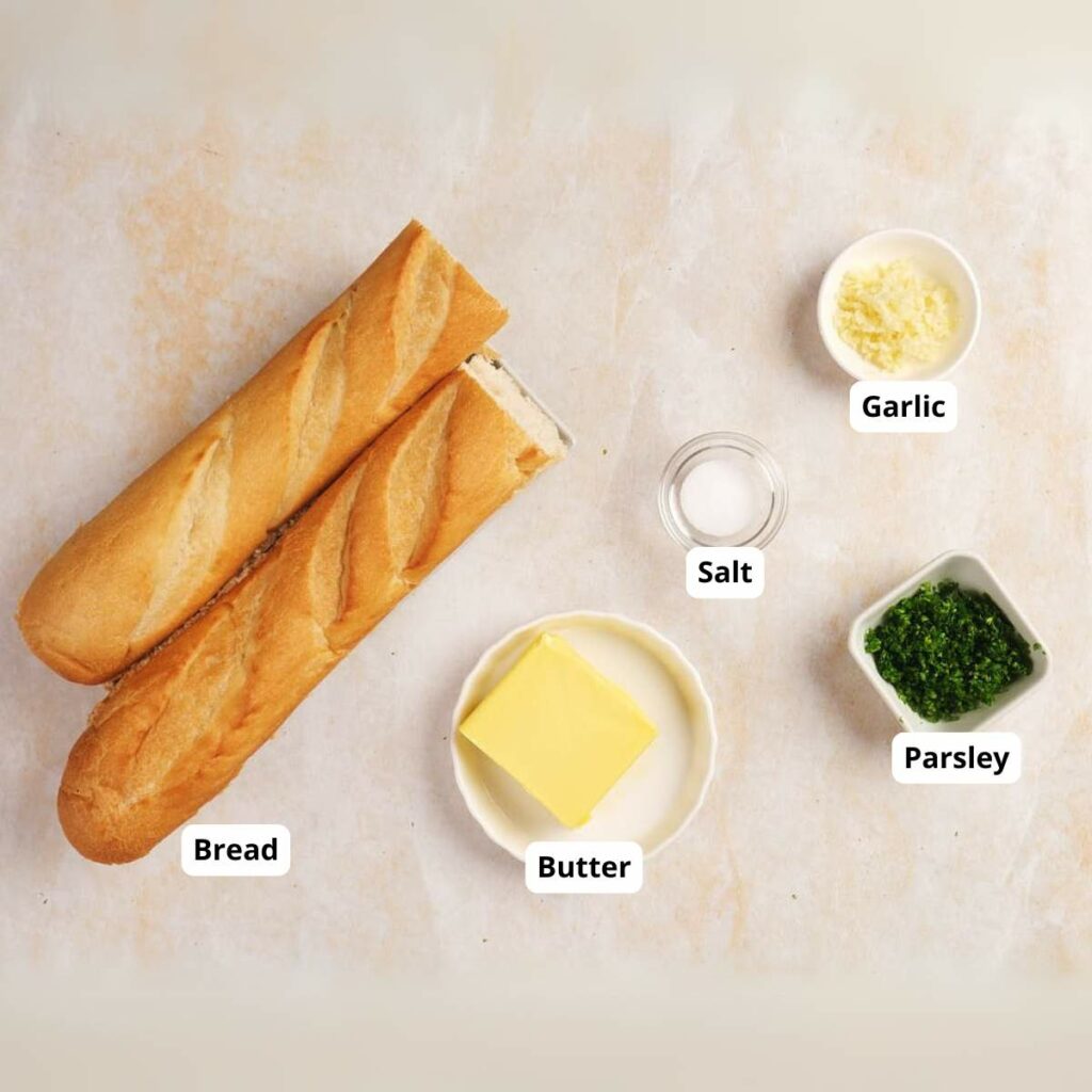 Two baguettes, a butter pat, garlic, salt, and parsley arranged on a light surface, each with a label identifying the ingredient.