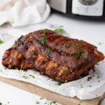 Baby back ribs garnished with chopped herbs sits on parchment paper atop a wooden cutting board.