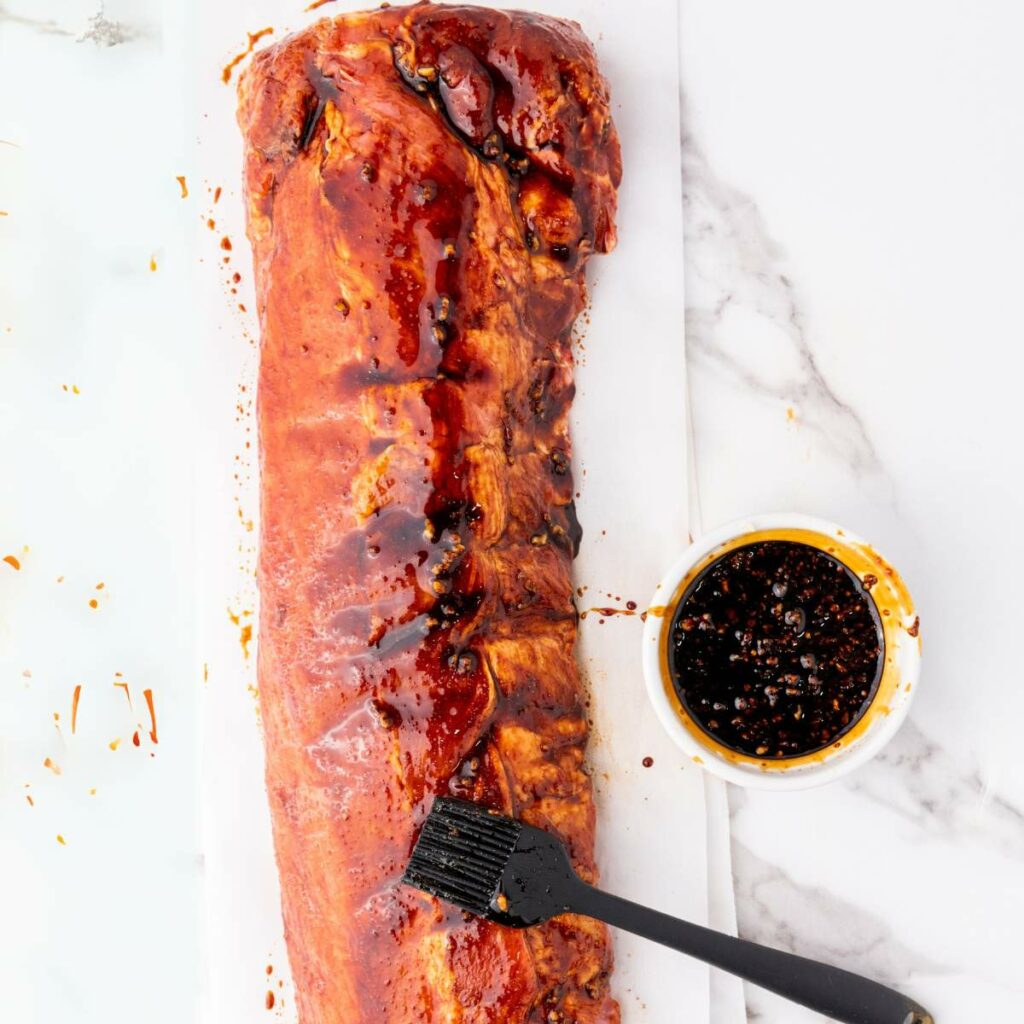 A large rack of ribs is being brushed with a dark sauce next to a bowl of marinade on a white marble surface.