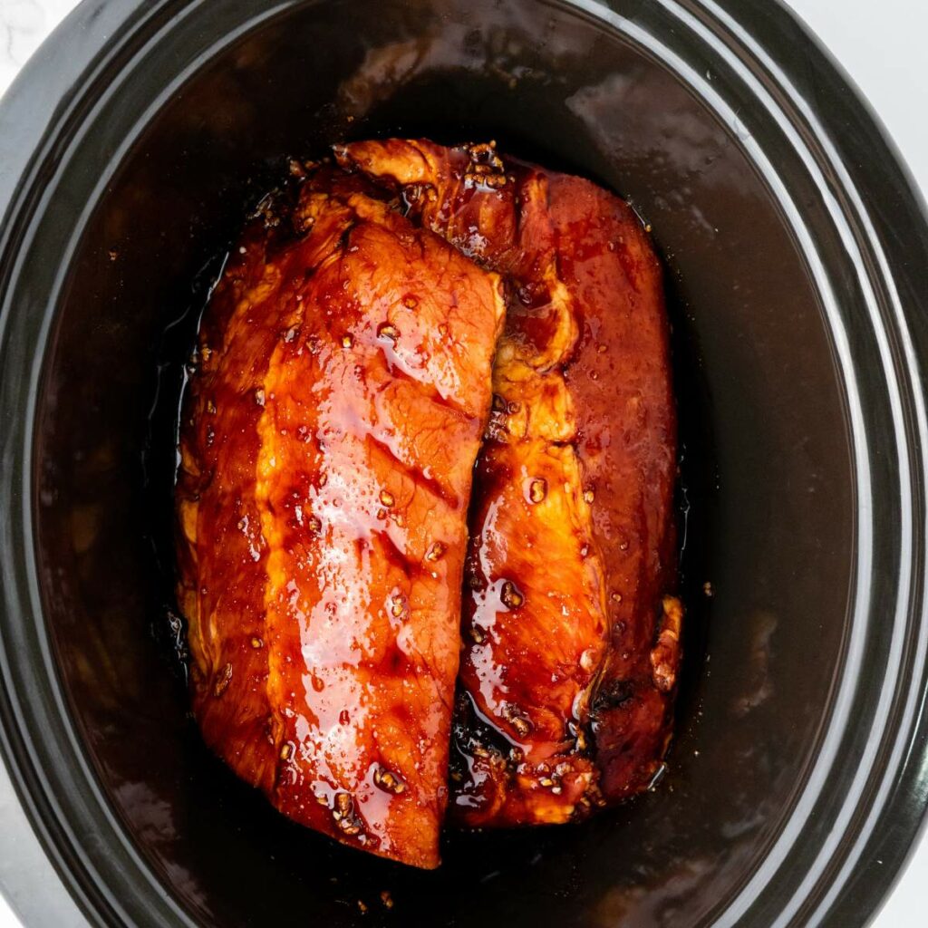 Two pieces of glazed meat are cooking inside a black slow cooker, coated in a shiny, dark sauce.