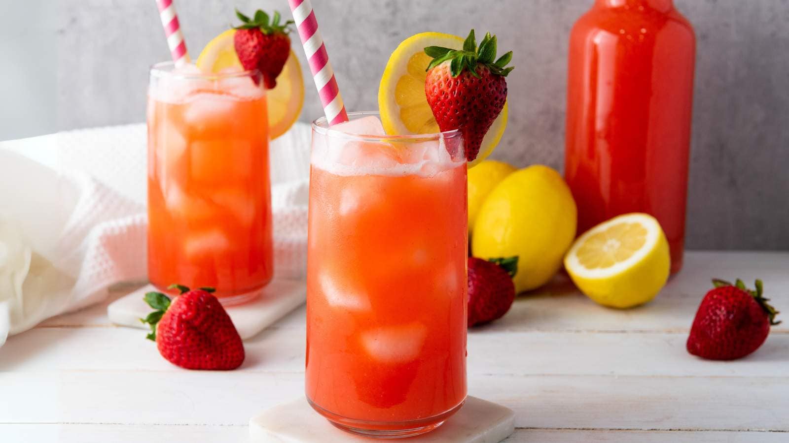 Two glasses of iced strawberry lemonade with striped straws, garnished with strawberries and lemon slices.
