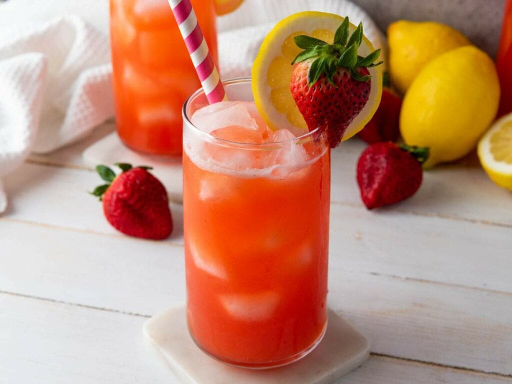 A glass of iced strawberry lemonade with a pink striped straw, garnished with a lemon slice and whole strawberry.