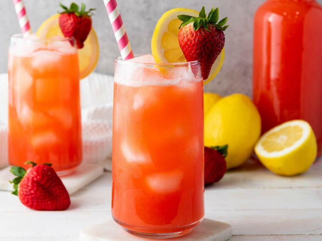 Two glasses of iced strawberry lemonade with pink-striped straws, garnished with lemon slices and strawberries.