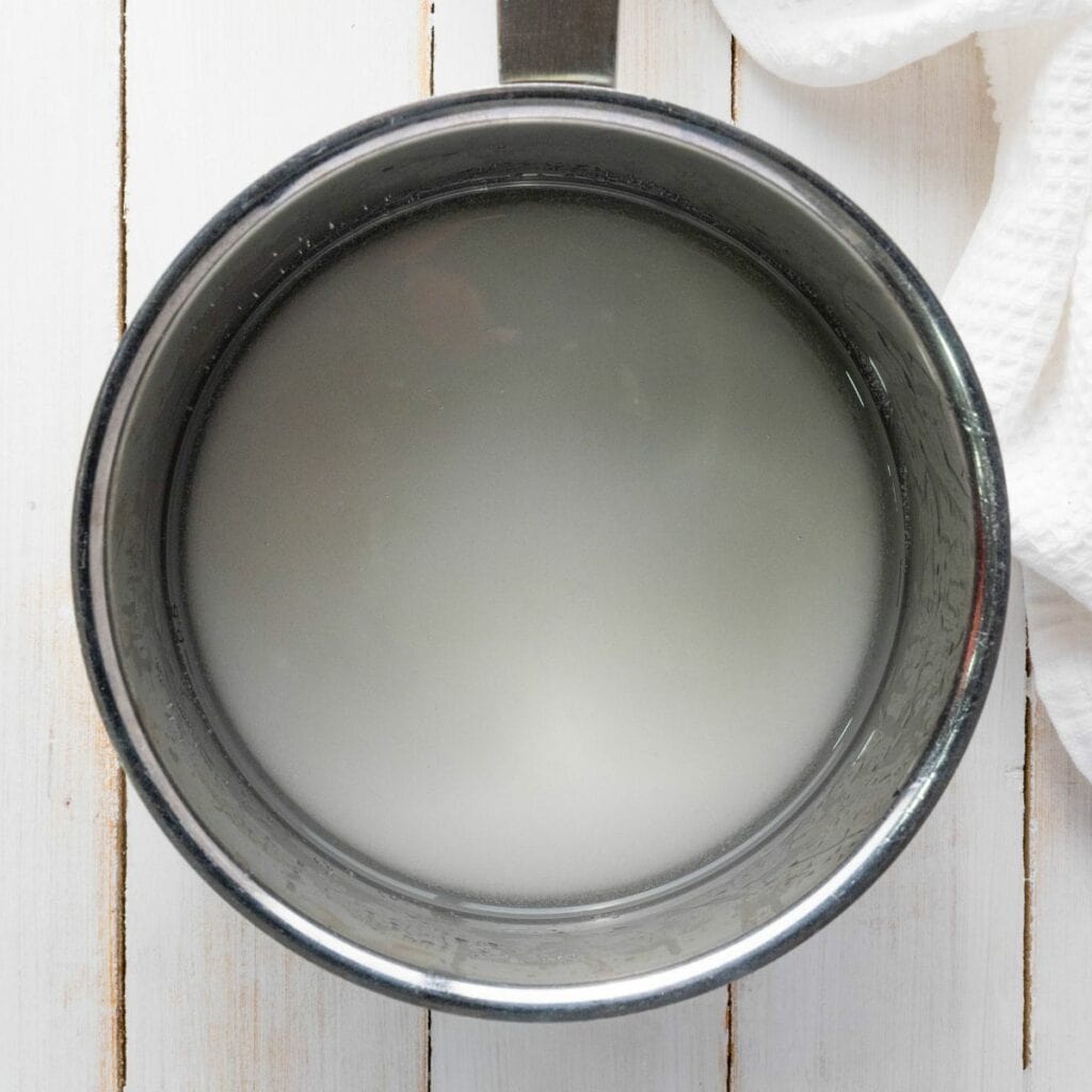 A stainless steel pot filled with cloudy water sits on a white wooden surface next to a white textured cloth.