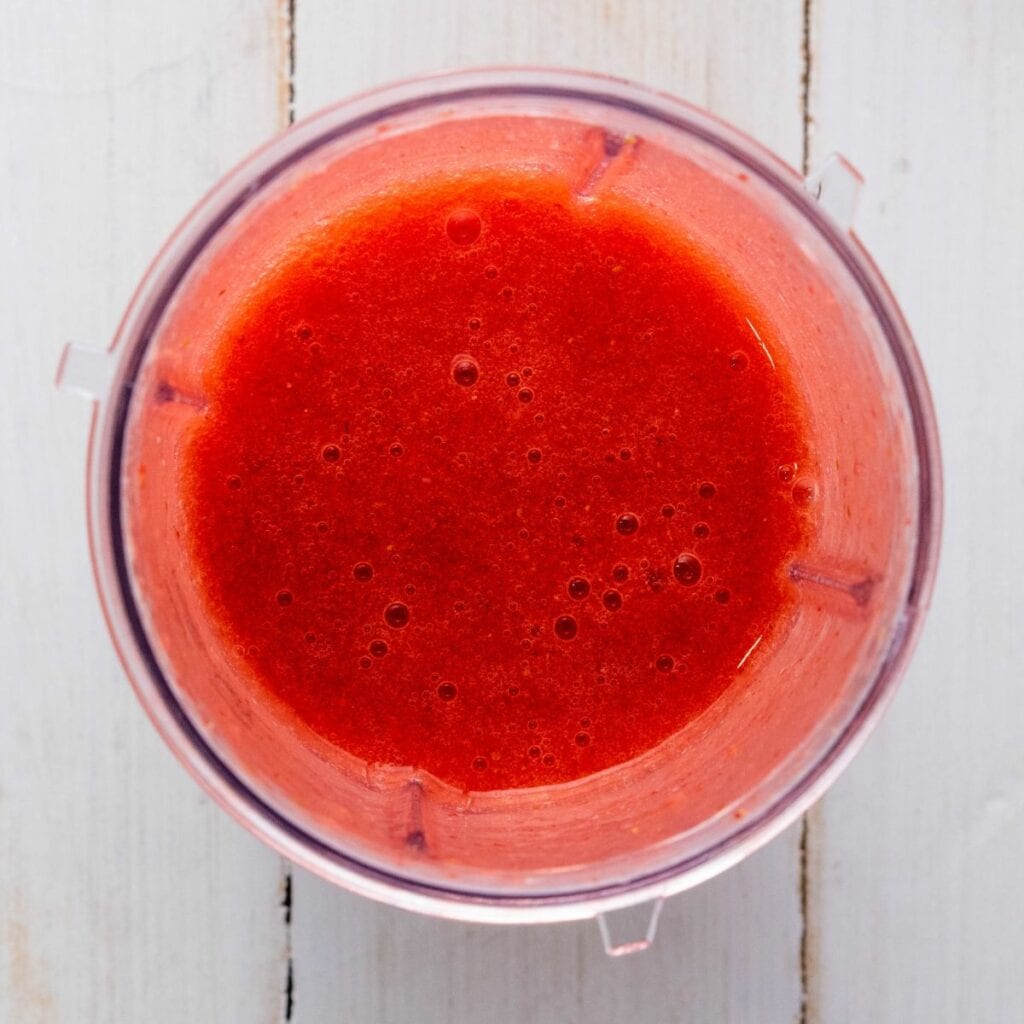 A blender cup filled with smooth, bright red liquid puree sits on a white wooden surface.