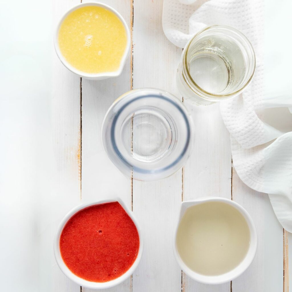Four containers with different liquids—yellow, red, clear, and pale yellow—are arranged on a white wooden surface beside a white towel.