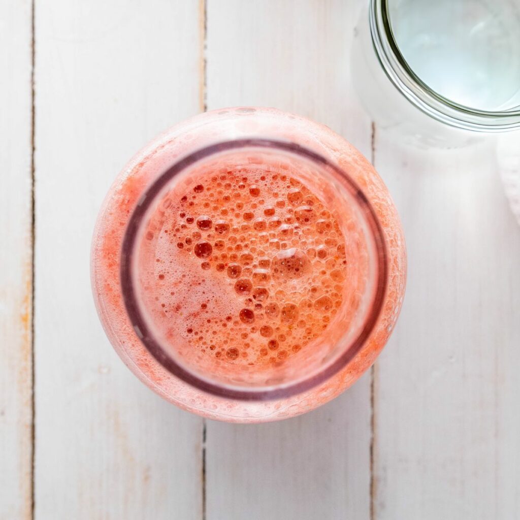 A top-down view of a glass jar filled with pink, frothy liquid on a light wooden surface, with part of an empty glass nearby.
