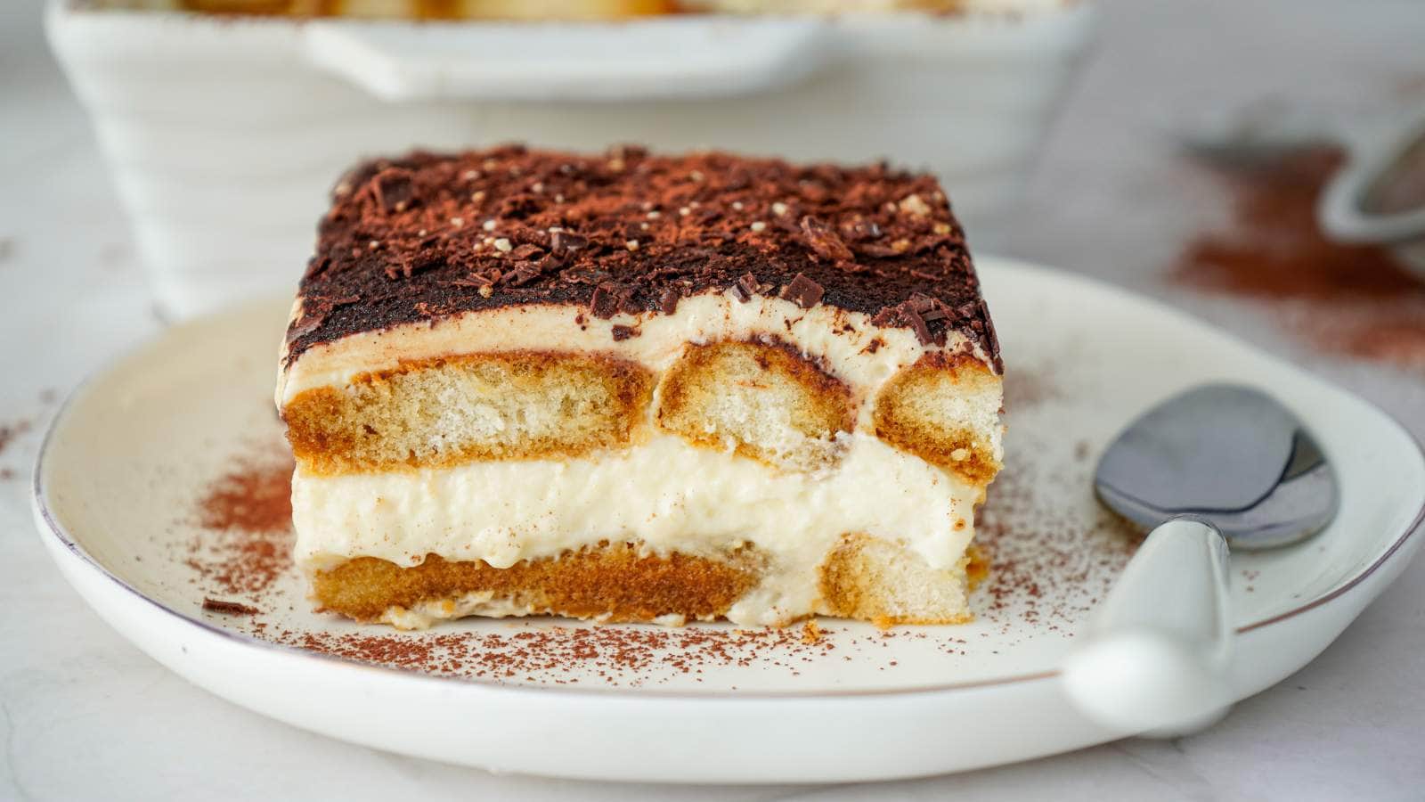 A slice of tiramisu with layers of cream and coffee-soaked ladyfingers, topped with chocolate shavings, served on a white plate with a spoon.
