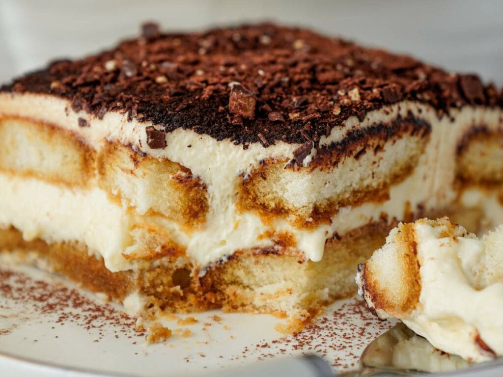 A close-up of a slice of tiramisu showing layers of mascarpone cream, soaked ladyfingers, and cocoa powder, with a spoonful taken out.