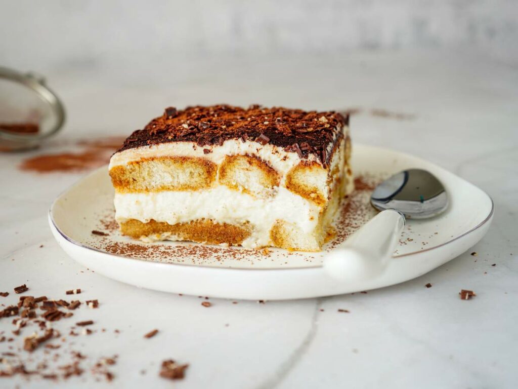 A slice of tiramisu topped with chocolate shavings on a white plate with a spoon, set on a marble surface.