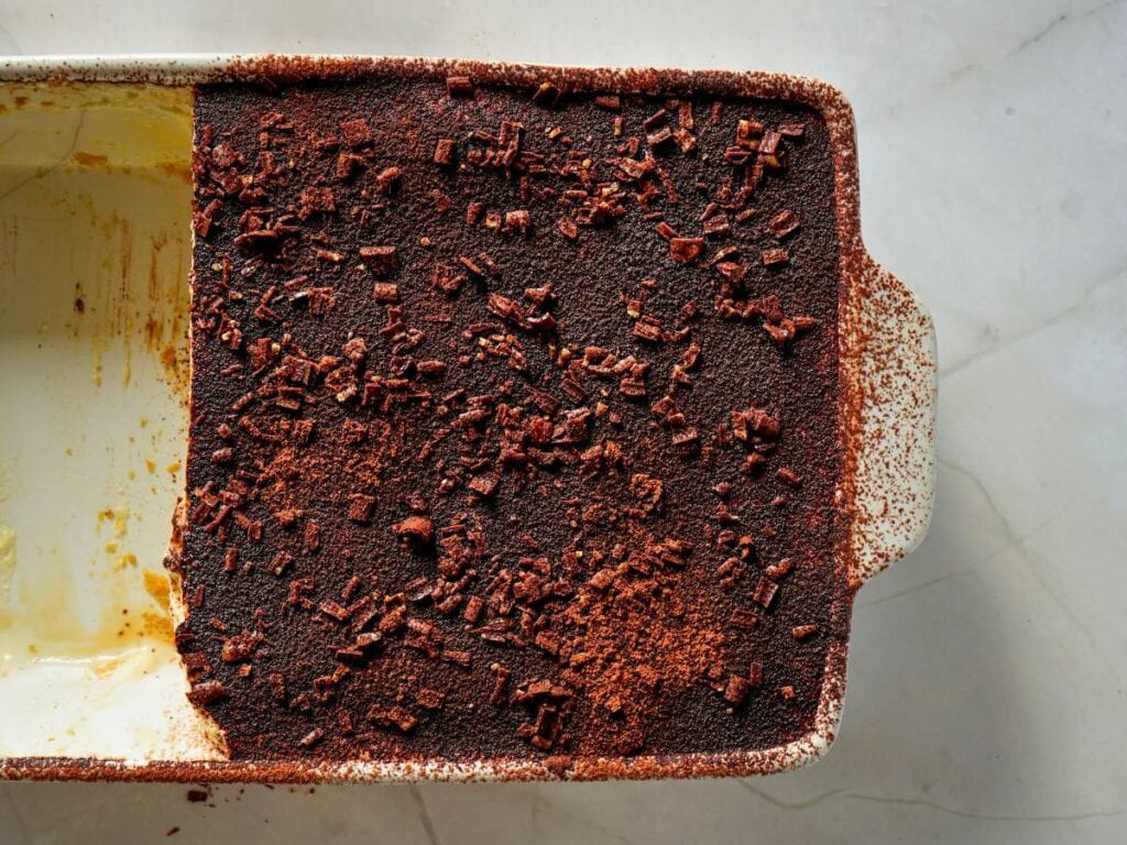 A rectangular dish of tiramisu with half of the dessert removed, showing a cocoa powder and chocolate topping on a marble surface.