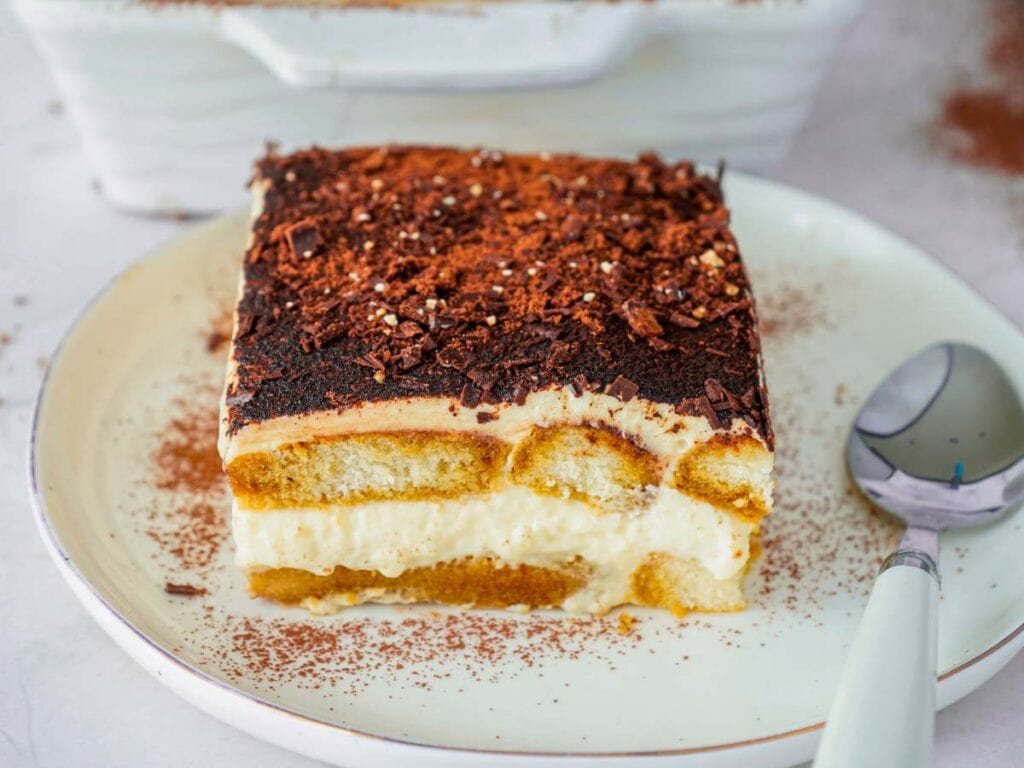 A slice of tiramisu on a white plate with a spoon beside it, dusted with cocoa powder and chocolate shavings.