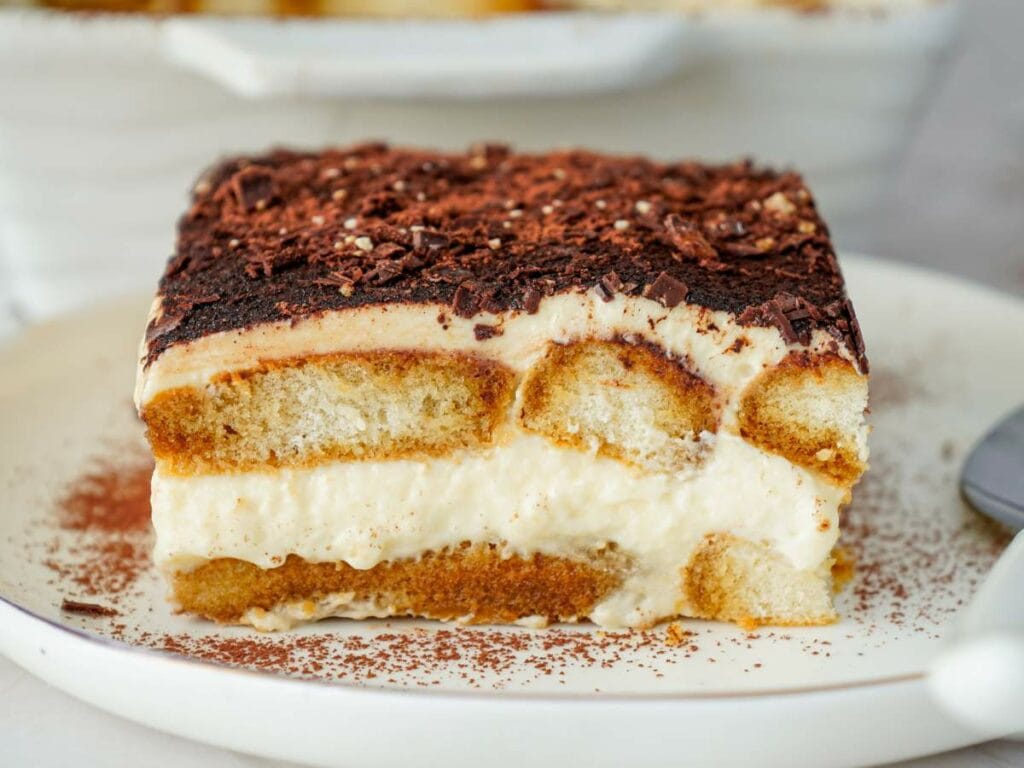 A close-up of a slice of tiramisu on a white plate, showing layers of cream, ladyfingers, and cocoa powder on top.