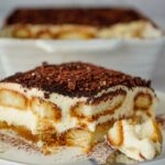 A close-up of a slice of tiramisu on a plate with a fork, showing layers of cream, soaked ladyfingers, and cocoa powder.