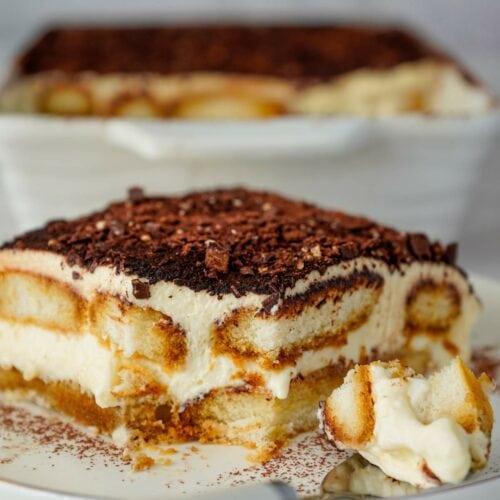 A close-up of a slice of tiramisu on a plate with a fork, showing layers of cream, soaked ladyfingers, and cocoa powder.