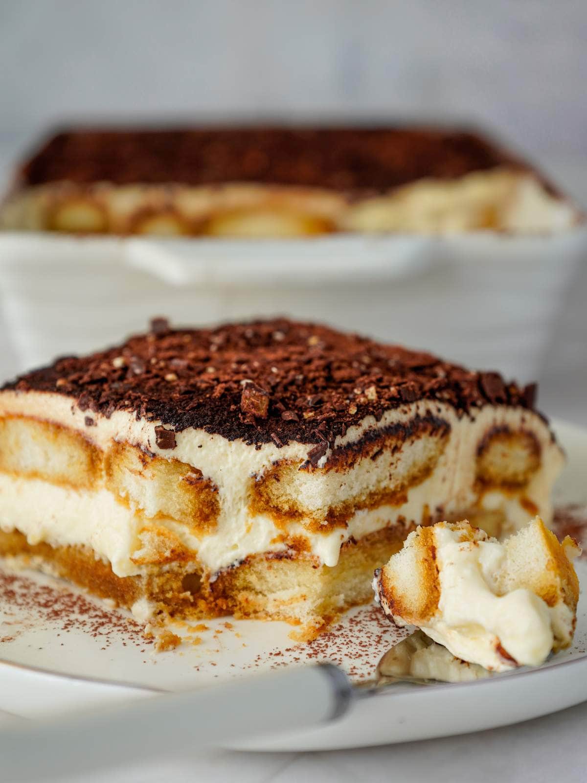 A close-up of a slice of tiramisu on a plate with a fork, showing layers of cream, soaked ladyfingers, and cocoa powder.