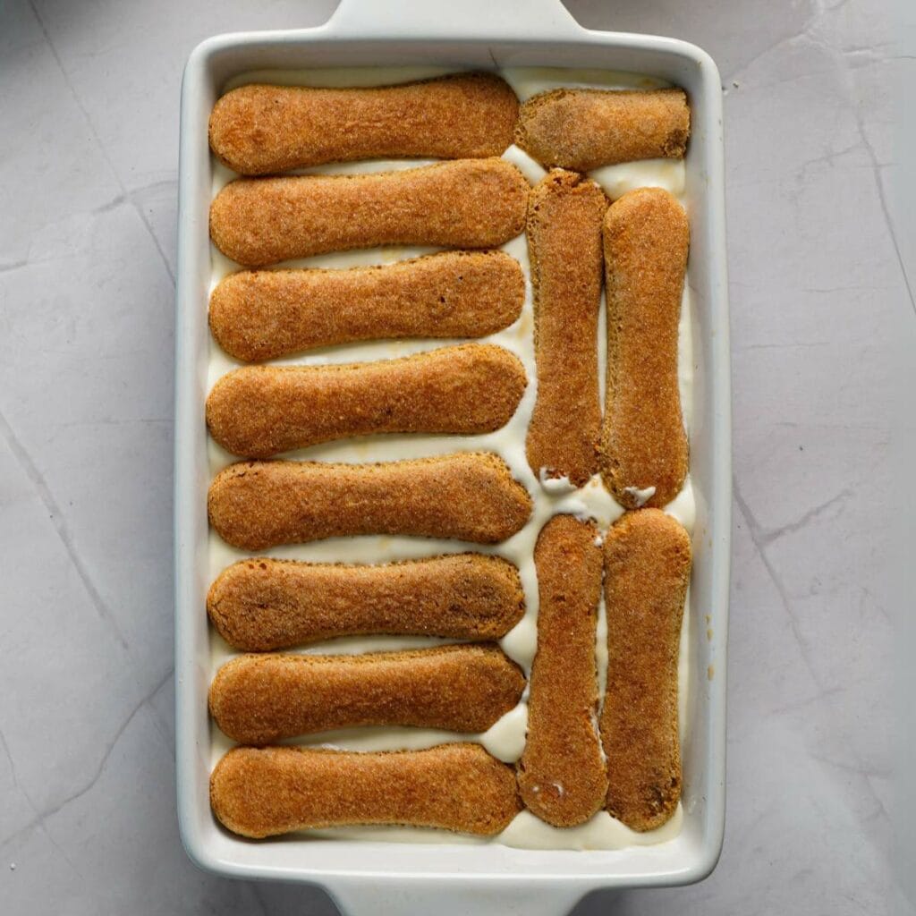 A rectangular dish with a layer of cream topped with ladyfinger biscuits arranged in rows, some overlapping, on a gray surface.