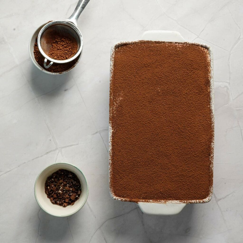 A rectangular dish of tiramisu topped with cocoa powder sits on a marble surface.