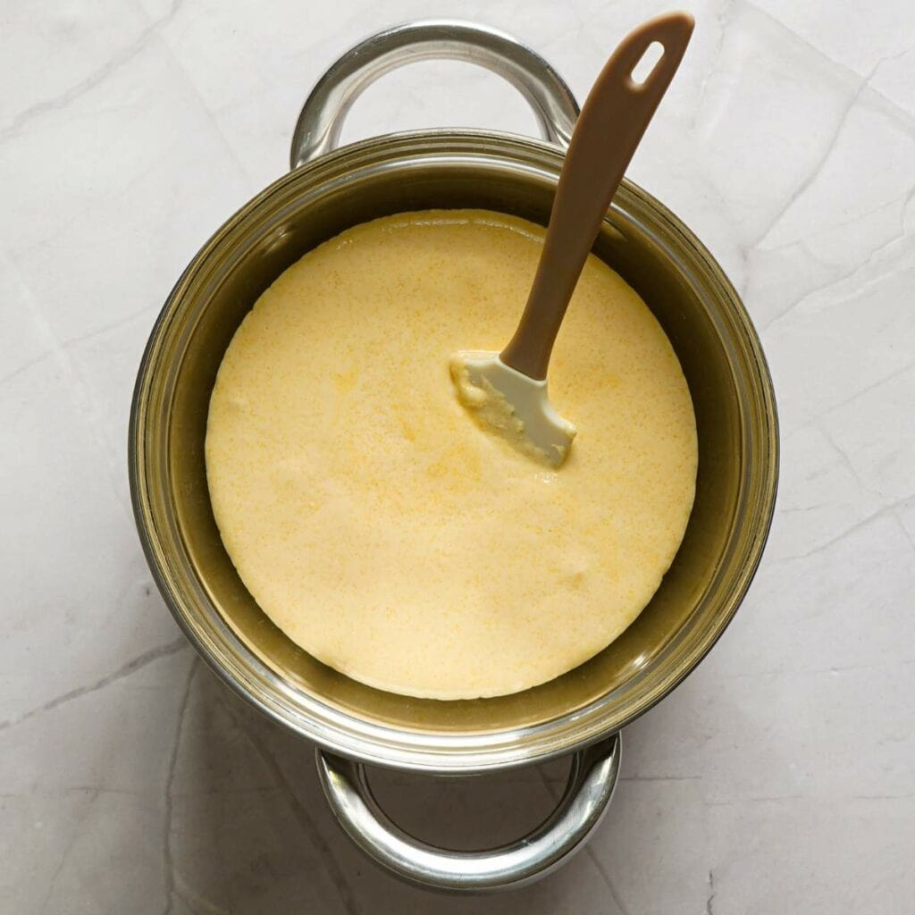 A metal pot containing a smooth, yellow, creamy mixture with a spatula resting on top, placed on a light marble surface.
