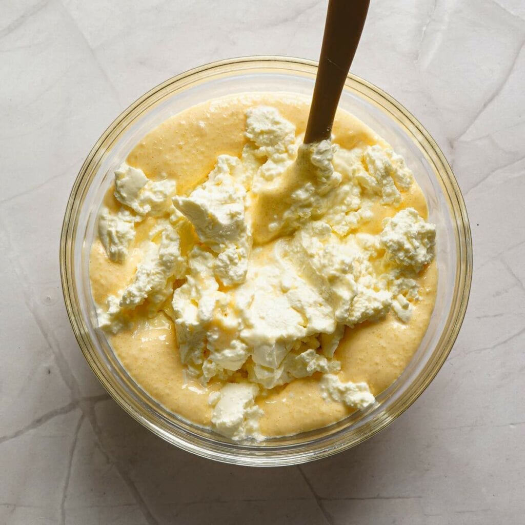 A bowl of yellow batter mixed with white chunks, possibly cottage cheese or ricotta, with a spoon on a light-colored marble surface.