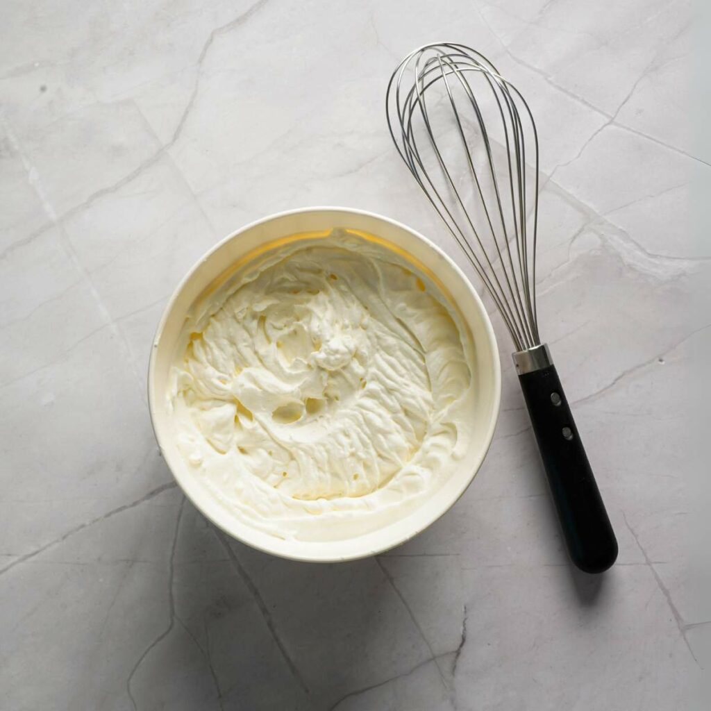 A bowl of whipped cream sits next to a metal whisk on a light gray marble surface.