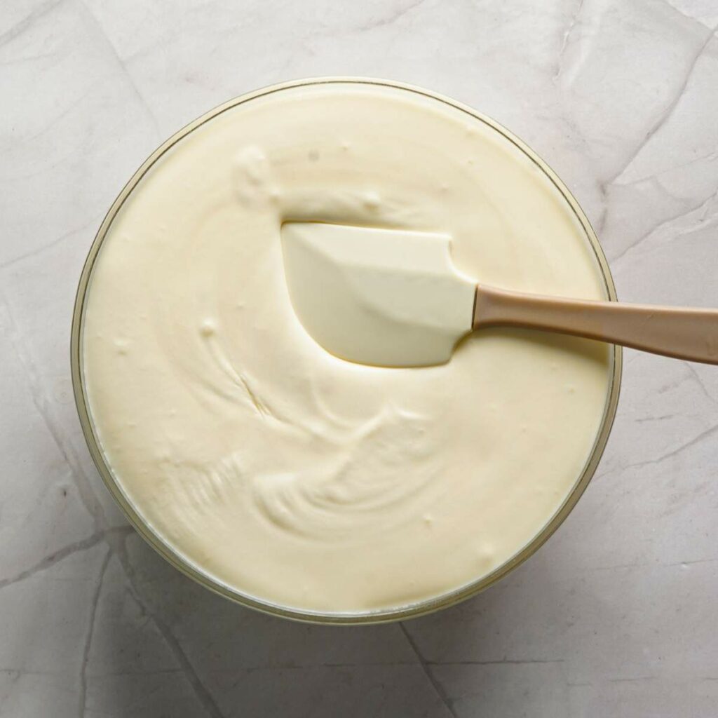 A glass bowl filled with creamy batter or mixture is being stirred with a silicone spatula on a light marble surface.