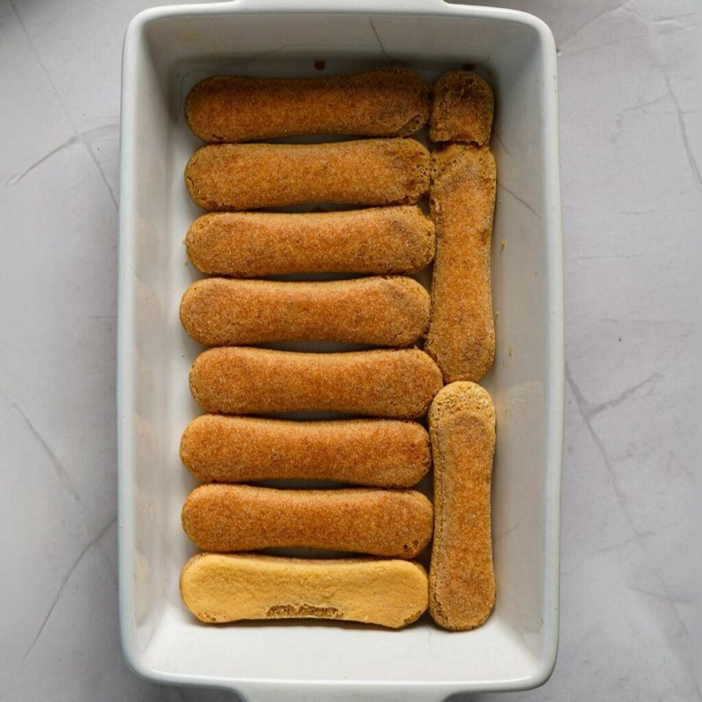 A rectangular dish with two rows of ladyfinger biscuits arranged neatly on the bottom, ready for the next step in preparation.