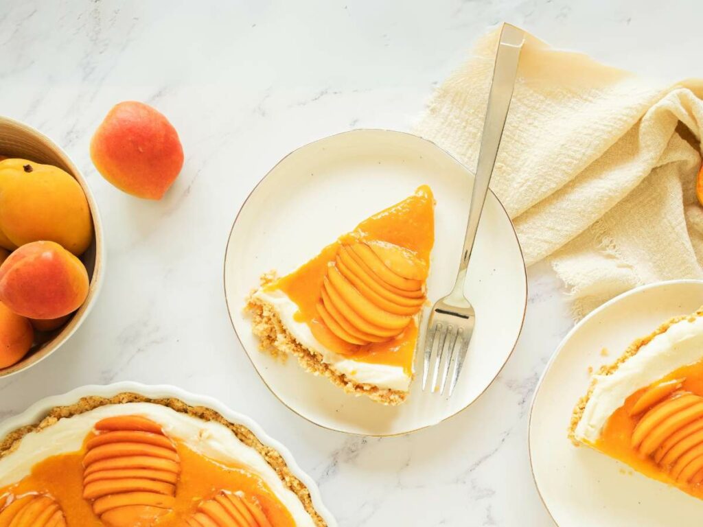 A slice of apricot cheesecake with a fork on a plate, a bowl of whole apricots, and the remaining tart on a marble surface.