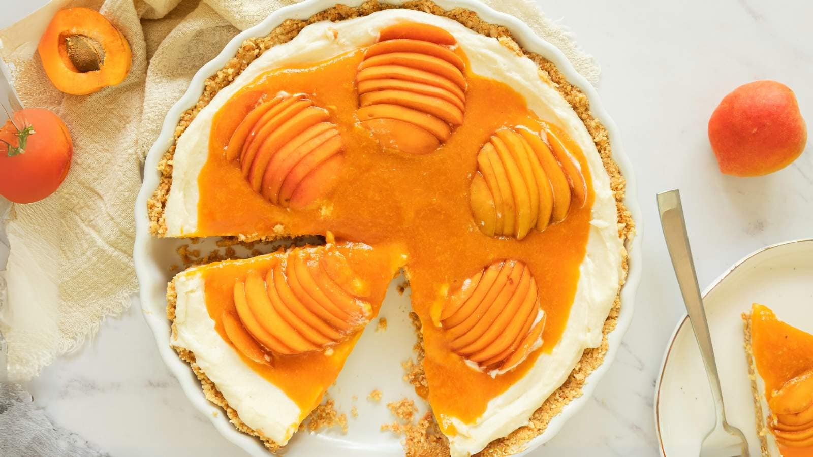 Apricot cheesecake with a creamy base, topped with sliced fresh apricots and apricot glaze, missing one slice on a white plate nearby.