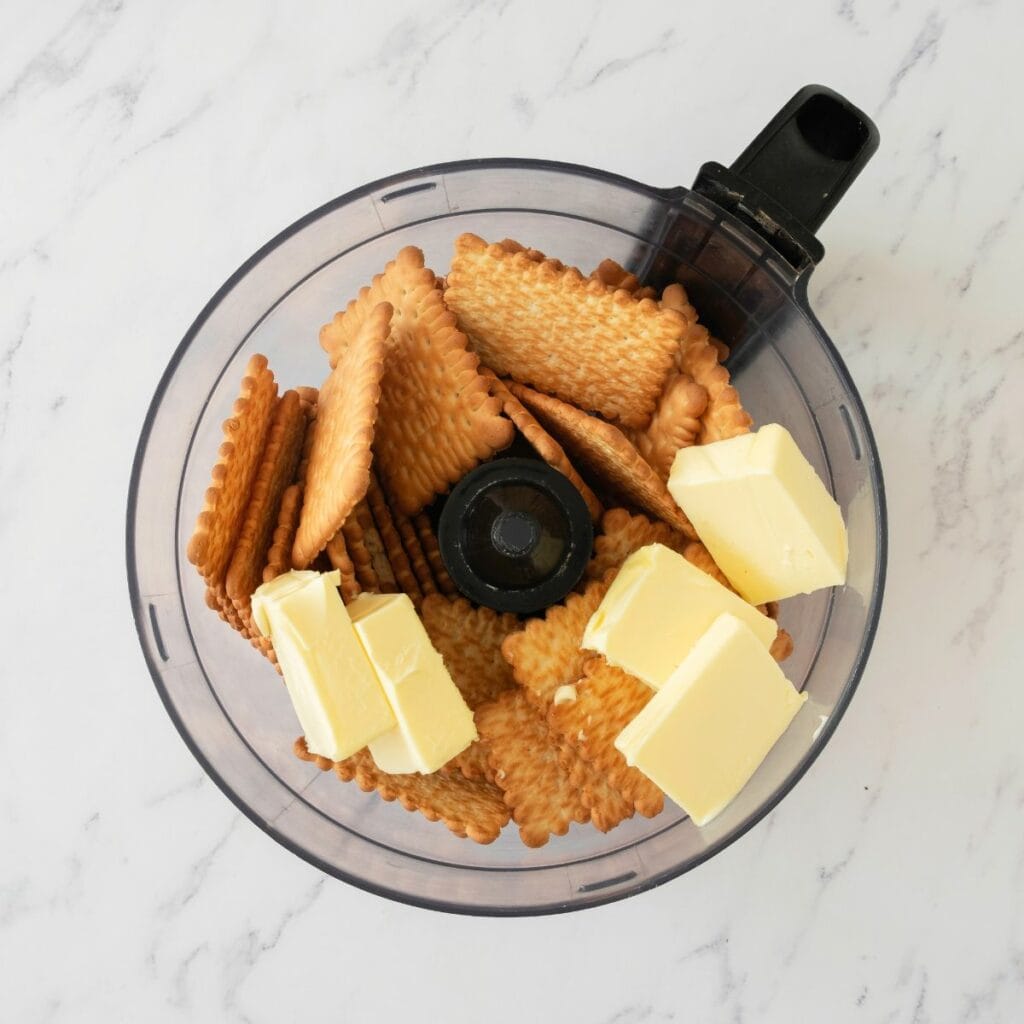 A food processor bowl containing rectangular biscuits and several chunks of butter on a white marble surface.