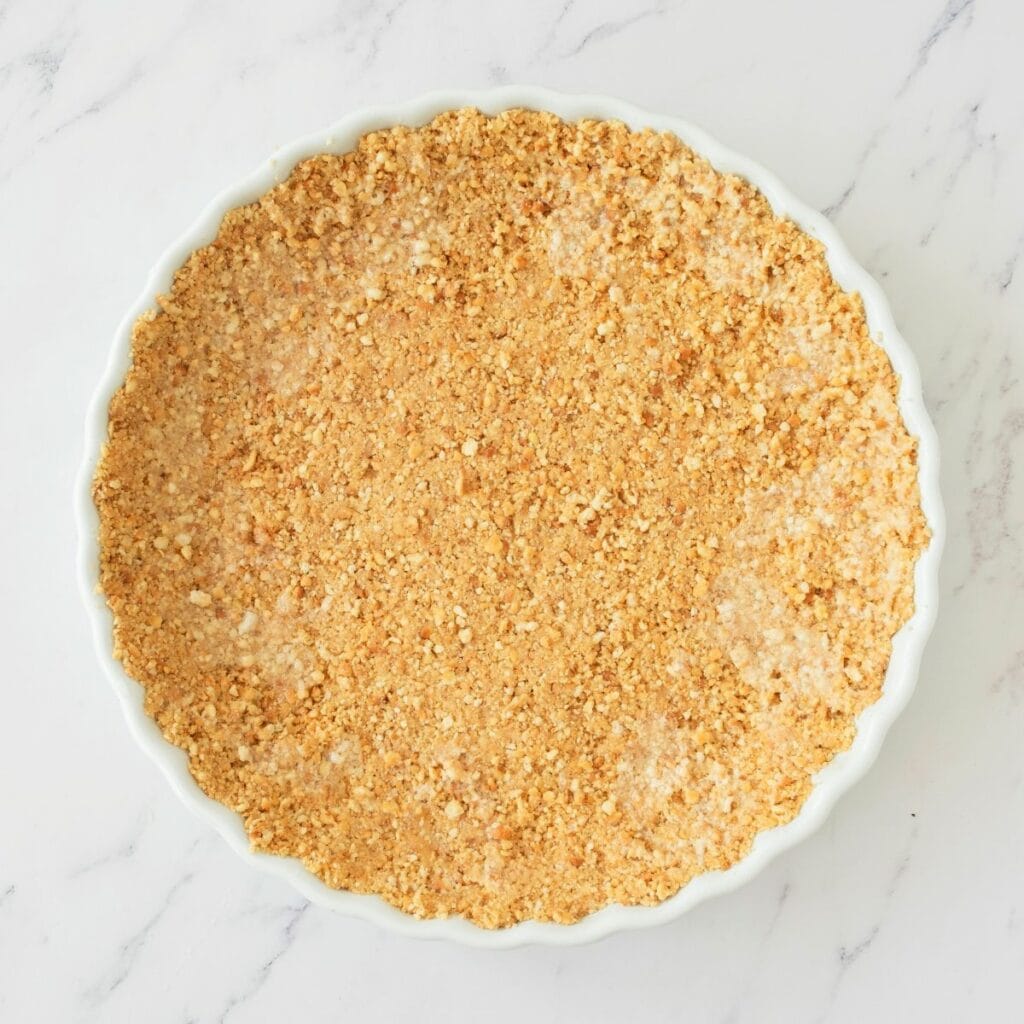 Round white dish filled with an even layer of crumbly, light brown pie crust on a white marble surface.