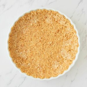 Round white dish filled with an even layer of crumbly, light brown pie crust on a white marble surface.