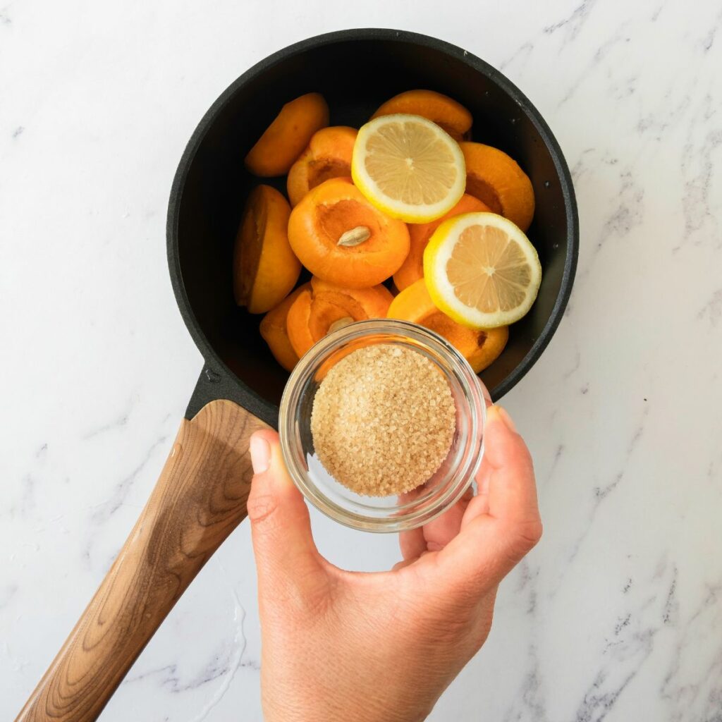 A hand holds a small glass bowl of brown sugar over a saucepan filled with sliced apricots and lemon rounds on a white marble surface.