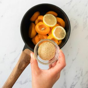 A hand holds a small glass bowl of brown sugar over a saucepan filled with sliced apricots and lemon rounds on a white marble surface.