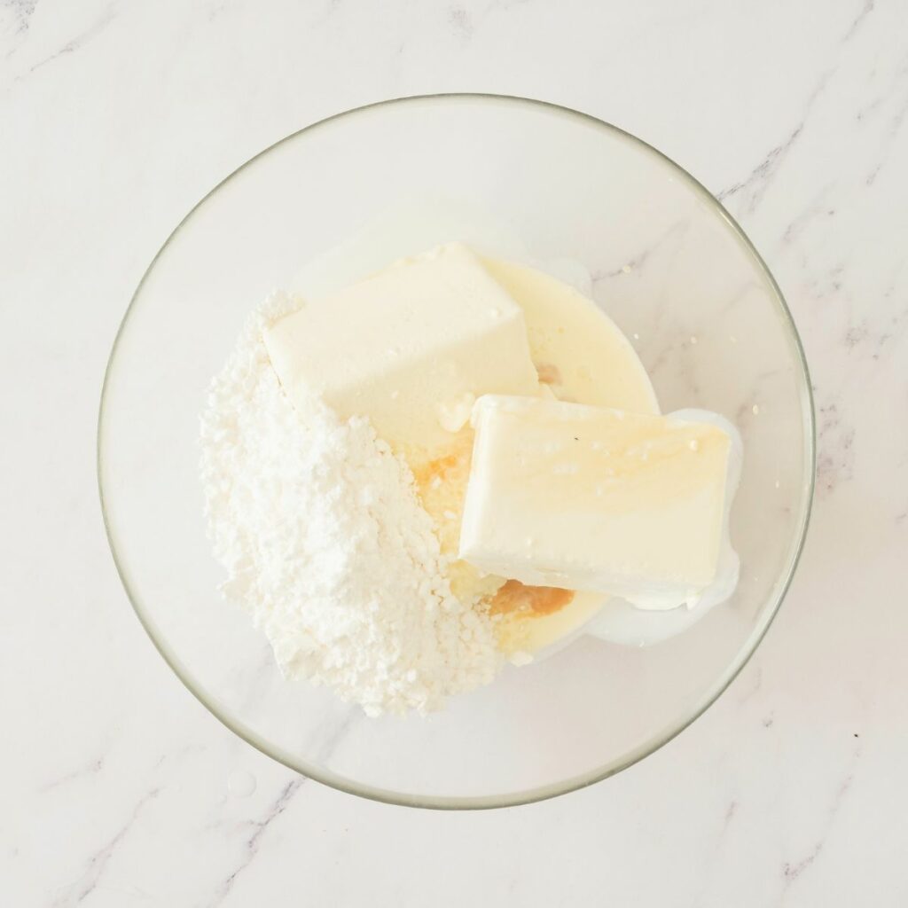 A glass bowl containing cream cheese, butter, powdered sugar, and a liquid ingredient on a white marble surface.